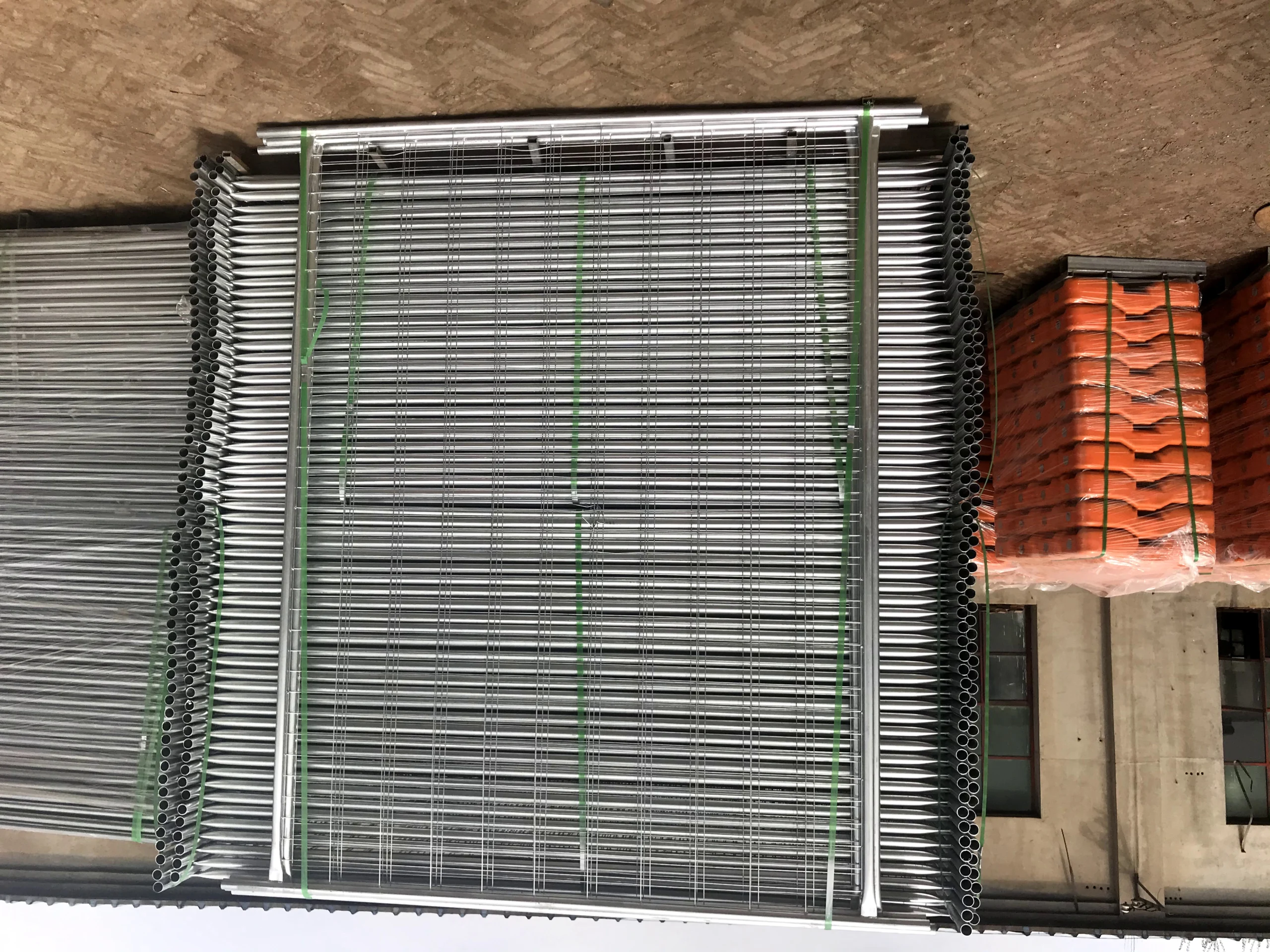 A warehouse displaying bulk stacks of hot-dipped galvanized temporary fence panels and bright orange plastic temporary fence feet, ready for export from Anping Deban Metal Wire Mesh Products Co., Ltd. As the sole supplier in Anping with its own plastic feet machine, DB Fencing offers comprehensive temporary fencing systems meeting Australian Standards for construction and event management.