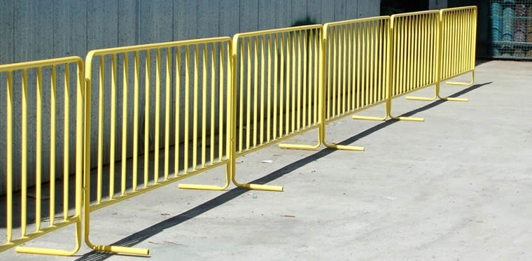 A long line of bright yellow metal crowd control barriers, manufactured by DB Fencing, stands on a concrete surface, casting shadows under sunlight. These durable, hot-dipped galvanized barriers are ideal for event management companies and global fence distributors requiring crowd control barriers for events and public safety.