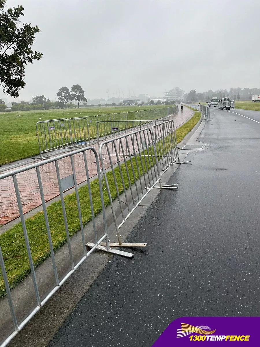 A long line of hot-dipped galvanized crowd control barriers from Anping Deban Metal Wire Mesh Products Co., Ltd (DB Fencing) defines a perimeter between a wet asphalt road and a grassy area, showcasing robust solutions for events and public safety. Designed to meet Australian standards and withstand harsh outdoor environments, these crowd control barriers are ideal for event management companies and global distributors in Australia, New Zealand, and beyond.
