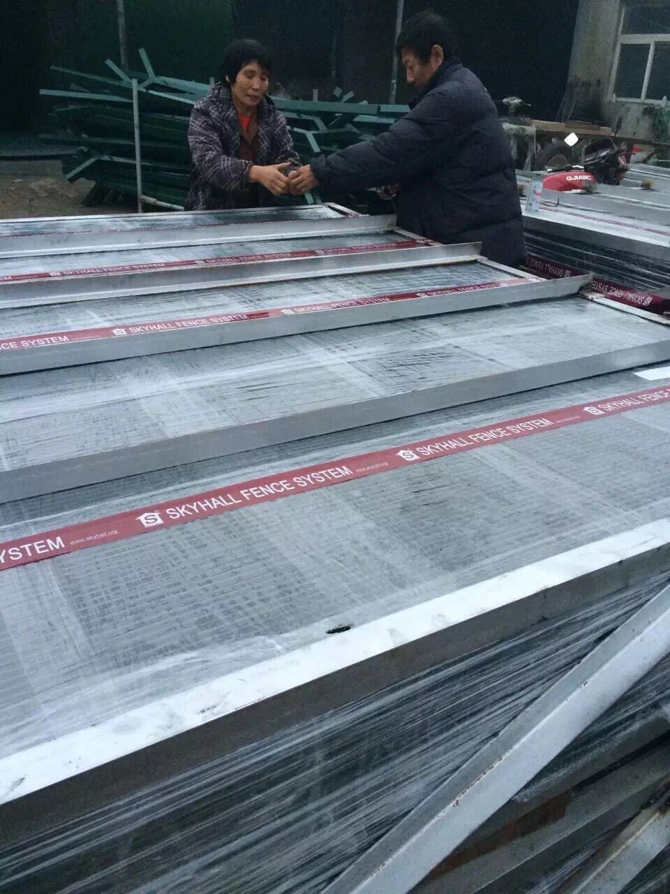 Workers inspecting large stacks of high-strength security fence panels at the DB Fencing manufacturing facility. The production line features hot-dipped galvanized materials designed for permanent perimeter security fencing solutions.