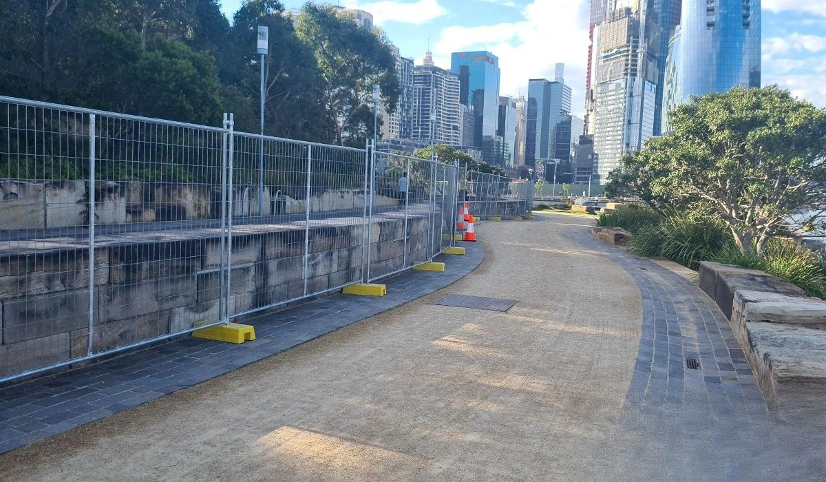 Robust hot-dipped galvanized temporary fencing systems, featuring DB Fencing's proprietary yellow plastic feet, line a waterfront pathway in a bustling urban construction zone. These AS 4687 compliant barriers provide essential site security and public safety in Australian environments.
